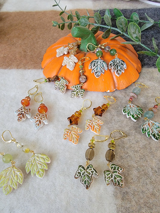 A styled flat-lay of handmade fall-themed drop earrings displayed around an orange pumpkin prop on a beige fabric surface. The earrings feature silver and green leaf charms, amber star beads, and green and orange beads.